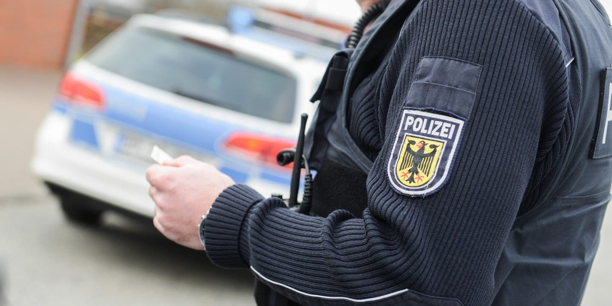 Polizeibeamter telefoniert, im Hintergrund Polizeiauto